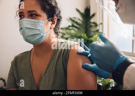 Nahaufnahme einer Frau, die eine Gesichtsmaske trägt und einen Coronavirus-Impfstoff auf ihren Arm geschossen hat. Frau, die sich zu Hause von einem Gesundheitshelfer impfen lassen. Stockfoto