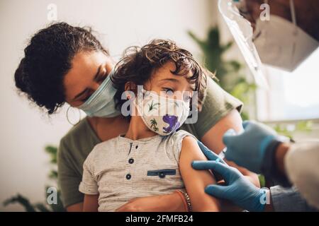 Kleiner Junge mit Gesichtsmaske, der den Impfstoff zu Hause nimmt. Kind mit Mutter, die zu Hause von einem Gesundheitshelfer kovideimpft wurde. Stockfoto