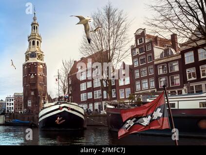 Stadtrundfahrt durch Amsterdam mit dem Montelbaanstoren-Turm in der Vorderansicht und Stadtbild am Kai des Kanals mit den niederländischen Häusern, Lastkähnen und Haus Stockfoto