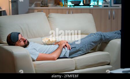 Der Mann schloss sein Auge und schlief auf der Couch im Wohnzimmer vor dem Fernseher ein, während die Fernsehunterhaltung LÄUFT. Entspannungskino nach einer harten Arbeitswoche Stockfoto
