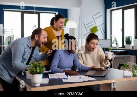 Das Startup Diversity Team freut sich, nach der Vertragsunterzeichnung mit einem wichtigen Kunden eine Partnerschaft mit Blick auf einen Laptop zu feiern. Fröhliches multiethnisches Geschäftsteam mit Laptop und Papieren, das sich für das Projekt begeistert. Stockfoto