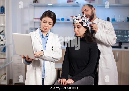 Facharzt für Neurologie, der am Laptop Notizen macht und die Symptome des Patienten fragt, indem er das High-Tech-eeg-Headset anpasst. Doctor Researcher Controlling EEG Headset Analyse Hirnfunktionen und Gesundheitszustand. Stockfoto