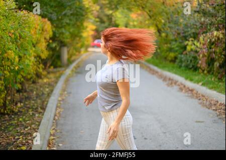 Portrait schöne rothaarige Frau in Brille und einem grauen T-Shirt geht im Park spazieren und flirtet. Ein glückliches Mädchen dreht sich um und winkt ihr Haar auf einem warmen Stockfoto