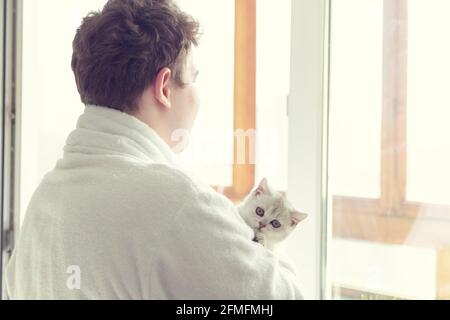 Der junge Kaukasusmann trinkt Kaffee in der Nähe des Fensters. Er hält eine Katze. Am frühen Morgen, Sonnenschein. Stockfoto