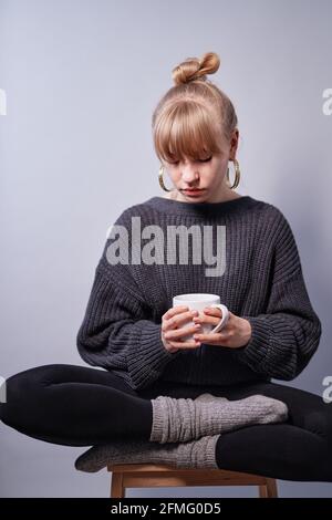 Kaukasische blonde Frau trägt schwarzen Pullover und schöne Ohrringe sitzen und halten Tasse Kaffee oder Tee. Romantische Stimmung oder Morgenkaffee-Konzept. Hochwertige vertikale Fotos Stockfoto