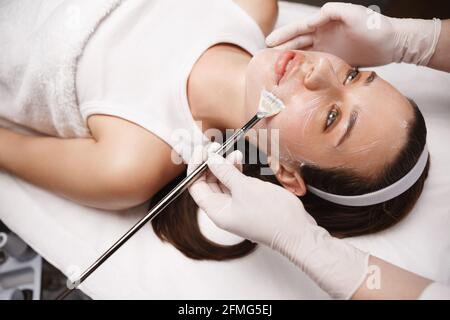 Junge Frau entspannt sich in der Schönheitsklinik. Kosmetikerin Hände in sterilen Handschuhen Pinsel Galvanic Gel, bereiten Frauen Haut für die Gesichtsbehandlung bei der Kosmetik Stockfoto