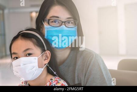 Portrait schöne asiatische Mutter und kleine Tochter tragen medizinische Maske, sitzen und warten zusammen auf einen Impfstoff in einem Krankenhaus, schwarze Haare, Augen l Stockfoto