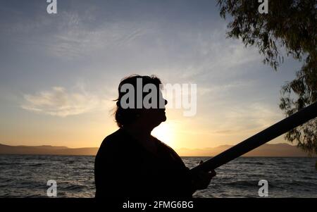 Silhouette bei Sonnenuntergang einer 62-jährigen Frau, der See von Galilee, die Unteren Galiläischen Berge und der bunte wolkige Himmel über ihnen sind hinter. Stockfoto