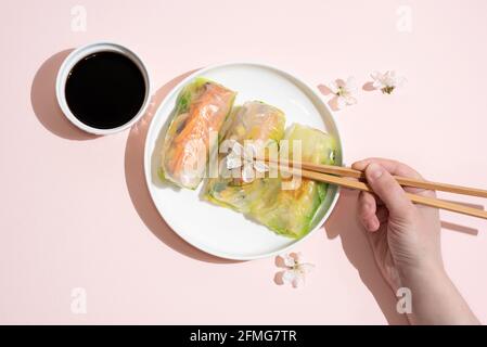 Weibliche Hand hält Essstäbchen und Teller mit Frühlingsrollen und Sojasauce auf rosa Hintergrund, asiatische Lebensmittel, Nahaufnahme. Stockfoto