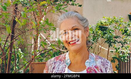 Reife Frau lächelt im Hinterhof Haus mit Sonne und Pflanzen. Weiß grauhaarige Frau zu Hause. Stockfoto