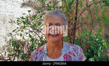 Reife Frau lächelt im Hinterhof Haus mit Sonne und Pflanzen. Weiß grauhaarige Frau zu Hause. Stockfoto