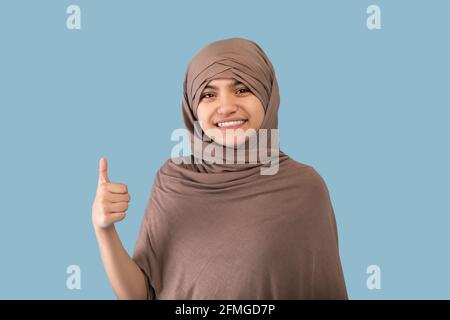 Positive lächelnde muslimische Teenager-Mädchen in Hijab zeigt Daumen nach oben Geste über blauen Studiohintergrund Stockfoto