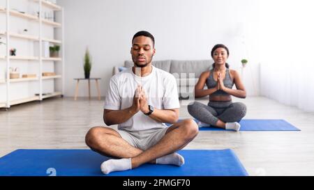 Schwarzer Kerl meditiert mit geschlossenen Augen, sitzt in Lotuspose, macht Namaste-Geste, praktiziert Yoga mit seiner Freundin Stockfoto