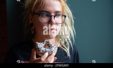 Ein ungewöhnliches Mädchen mit Dreadlocks auf dem Kopf und einem Piercing isst in einem Café einen Donut. Eine junge Frau in Gläsern frühstückt an einer grauen Wand Stockfoto