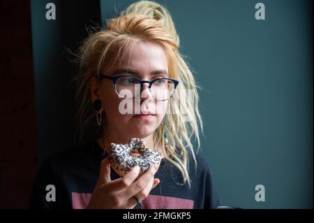 Ein ungewöhnliches Mädchen mit Dreadlocks auf dem Kopf und einem Piercing isst in einem Café einen Donut. Eine junge Frau in Gläsern frühstückt an einer grauen Wand Stockfoto