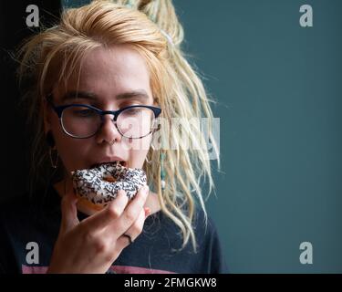 Ein ungewöhnliches Mädchen mit Dreadlocks auf dem Kopf und einem Piercing isst in einem Café einen Donut. Eine junge Frau in Gläsern frühstückt an einer grauen Wand Stockfoto