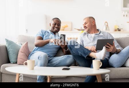 Freunde sitzen auf dem Sofa mit modernen Gadgets, Mann zeigt Witz am Telefon Stockfoto