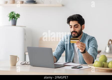 Araber, der online mit Laptop und Kreditkarte bezahlt, zu Hause in Kithcen sitzt, leerer Raum Stockfoto