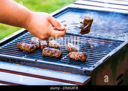 Eine Nahaufnahme der Hand, die Fleischbrötchen, genannt Mici oder, zubereitet Mititei auf Grill mit aufsteigendem Rauch Stockfoto