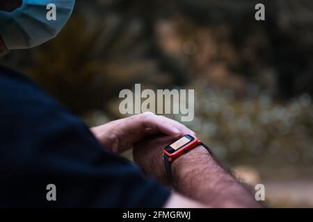 Ein Mann, der eine Maske trägt, interagiert und seine anschaut Smart-Watch, um seine Herzfrequenz und seine zurückgelegte Strecke zu sehen Stockfoto