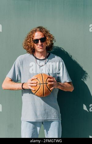 Vertikales Porträt eines dünnen langhaarigen kaukasischen jungen Mannes. Er hält einen Basketballball mit beiden Händen. Die Kamera ernst nehmen Stockfoto