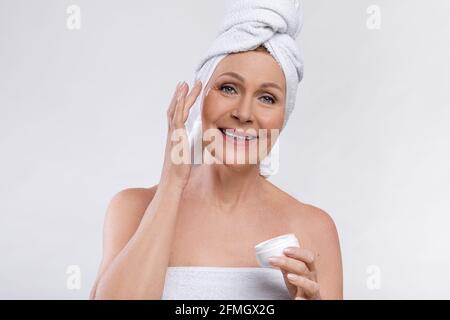 Schöne ältere Frau, die Gesichtscreme aus dem Glas aufträgt und Badetücher auf hellem Studiohintergrund trägt Stockfoto