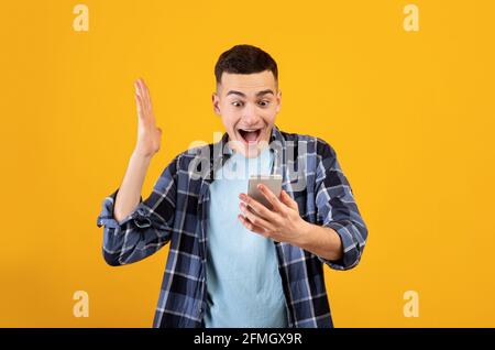 Aufgeregt Millennial Kerl in Casual Outfit Blick auf Smartphone, glücklich über seinen Sieg oder Erfolg auf orange Studio Hintergrund Stockfoto