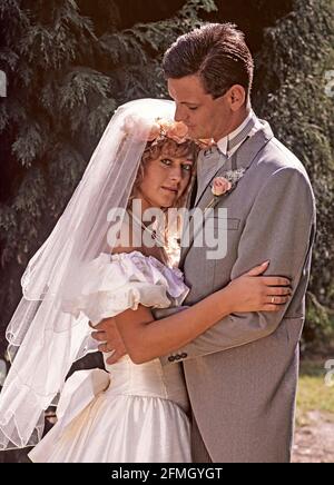 Historische Ansicht der Hochzeit in den 1980er Jahren des jungen Models freigegebenes Paar in romantischem Kuscheln Pose zusammen in Sonnenschein im Freien zu Andenken Fotos der Braut in 80er Jahre Mode Hochzeitskleid mit langem Schleier und Bräutigam trägt grauen Morgenanzug mit Knopfloch wie wir waren Archivbild in 1989 Essex England UK Stockfoto