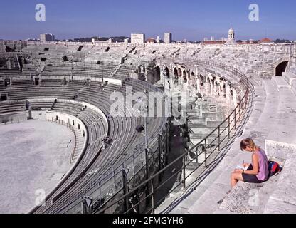 Historikal1990s Archiv Innenansicht der erhaltenen französischen Arena von Nîmes ein berühmtes Amphitheater aus dem Römischen Reich, das 1993 besichtigt wurde Von der höchsten Ebene mit jungen Frau Touristen sitzen auf Steinterrasse mit Blick auf die Innenansicht der 90er Jahre bis hin zur Funktionsfähigkeit Veranstaltungsarena und niedrigere Ebenen der Sitzgelegenheiten auf einem heißen Blauer Himmel Sonnenschein Tag in einem Archivbild Reise in Das Departement Gard der Region von Südfrankreich in Europa Stockfoto