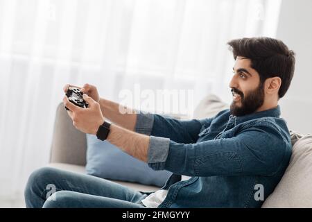 Zeitvertreib für Männer. Arabischer Kerl, der Online-Spiele spielt, Joystick Gamepad benutzt, auf der Couch im Wohnzimmer sitzt, Seitenansicht Stockfoto