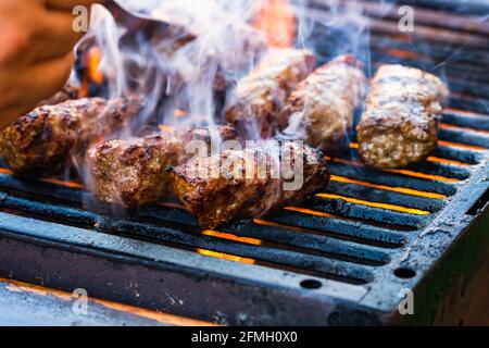 Eine Nahaufnahme der Hand, die Fleischbrötchen, genannt Mici oder, zubereitet Mititei auf Grill mit aufsteigendem Rauch Stockfoto