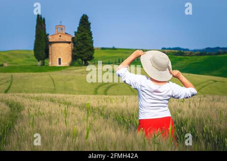 Schöne fröhliche Frau in rotem Rock und Strohhut genießen die Freiheit und die Aussicht. Landschaft mit Getreidefeldern und Vitaleta-Kapelle, Toskana, Stockfoto