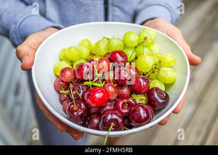 Nahaufnahme eines Mannes, der ganze Früchte mit Rot und Grün hält Weintrauben und Kirschen in Tellerschale außen mit Holzbokeh Hintergrund Stockfoto