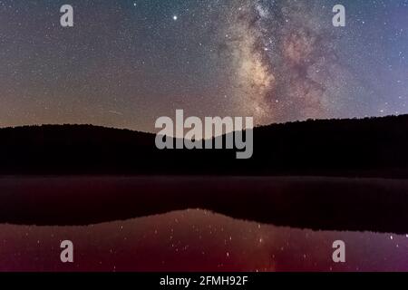 Dunkler Nachthimmel mit milchigen Sternen im Fichte Knauf See in West Virginia mit rot lila Farbe Wasser und Spiegelung der Landschaft der Galaxie Stockfoto