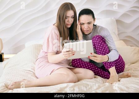 In Nachtkleidung gekleidete Frauen sehen sich auf dem Bildschirm eines elektronischen Tablets einen Film an, während sie im Schlafzimmer auf dem Bett sitzen. Stockfoto