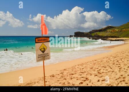 'Warnung: Gefährlicher Shorebreak'-Schild am Makapuu Beach mit vulkanischen Felsen in der Ferne, Waimanalo, Oahu, Hawaii, USA Stockfoto