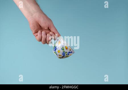 Hand und Tasse auf blauem Hintergrund. Mann hält weiße Tasse mit bemalten Blumen. Keramik Becher Tasse für Tee oder Kaffee hell dekoriert in blau und gelb c Stockfoto