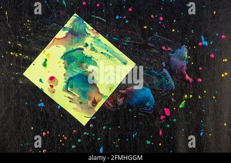 Chaotische Farbtropfen und ein Stück Papier auf schwarzer Textur. Gelbe, rote, blaue Punkte und Spritzer. Grünes quadratisches Blatt Papier mit Farben befleckt. Abst Stockfoto