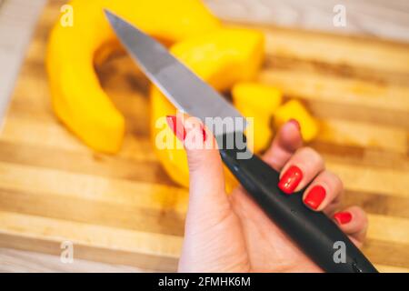 Weibliche Hand mit roter Maniküre hält Messer mit verwundeter Hand Auf dem Hintergrund des Schneidebretts mit Kürbis Stockfoto