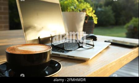 Wunderschöner, fauler, goldener Morgensonne auf einem Tisch im Freien mit heißem Kaffee, einem Laptop, Handy, Gläsern und einer Maus für Onli Stockfoto