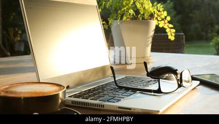 Schöner goldener Morgensonne auf einem Tisch im Freien mit heißem Kaffee, einem Laptop, Handy, Brille und Maus bereit für onlin Stockfoto