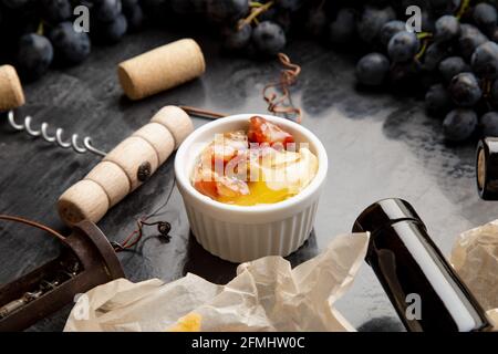 Honig im Glas auf Tisch aus dunklem Stein mit Weinkäse Korkenzieher in schwarzem saftigen Trauben Rahmen. Köstlicher gelber Bienenhonig gemischt mit Nüssen und getrockneten Früchten Stockfoto