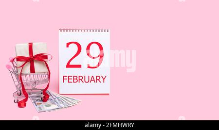 29. Tag im februar. Eine Geschenkbox in einem Einkaufswagen, Dollars und ein Kalender mit dem Datum des 29. februar auf einem rosa Hintergrund. Wintermonat, Tag von Stockfoto