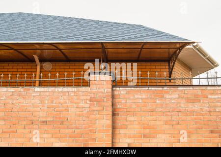 Die Mauer des Zauns besteht aus rotem Backstein, das Dach des Hauses ist von hinten sichtbar. Stockfoto