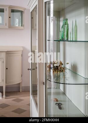 Weiße Schränke im alten Stil mit Glasregalen im Wohnzimmer. Grüne Glasflaschen und getrocknete Blumen zur Dekoration. Stockfoto