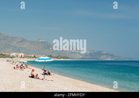 Nicht identifizierte Menschen am Strand in Giardini Naxos mit Taormina Stadt im Hintergrund, Sizilien Stockfoto