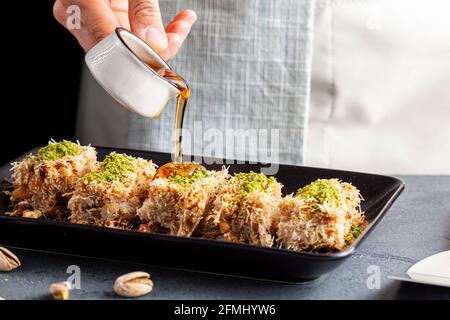 Ein kaukasischer Koch gießt dicken süßen Sirup aus einem Porzellanbecher auf Scheiben türkisches Kadayif-Dessert. Dies wird mit geschreddertem phyllo doug gemacht Stockfoto