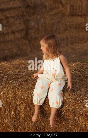 Unbeschwertes Kind in Overalls, das auf Strohballen bei Sonnenschein sitzt Tag auf dem Land Stockfoto