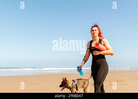 Lächelnde Sportlerin mit gerollter Matte und einer Flasche Wasser Schlendern Sie mit einem reinrassigen Hund an der sandigen Küste und schauen Sie nach Weg Stockfoto
