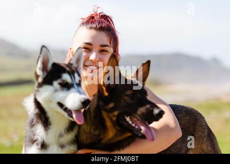 Inhalt weibliche Athletin mit Piercing umarmt sibirischen Husky und Deutsch Schäferhund beim Blick auf die Kamera im Sonnenlicht Stockfoto